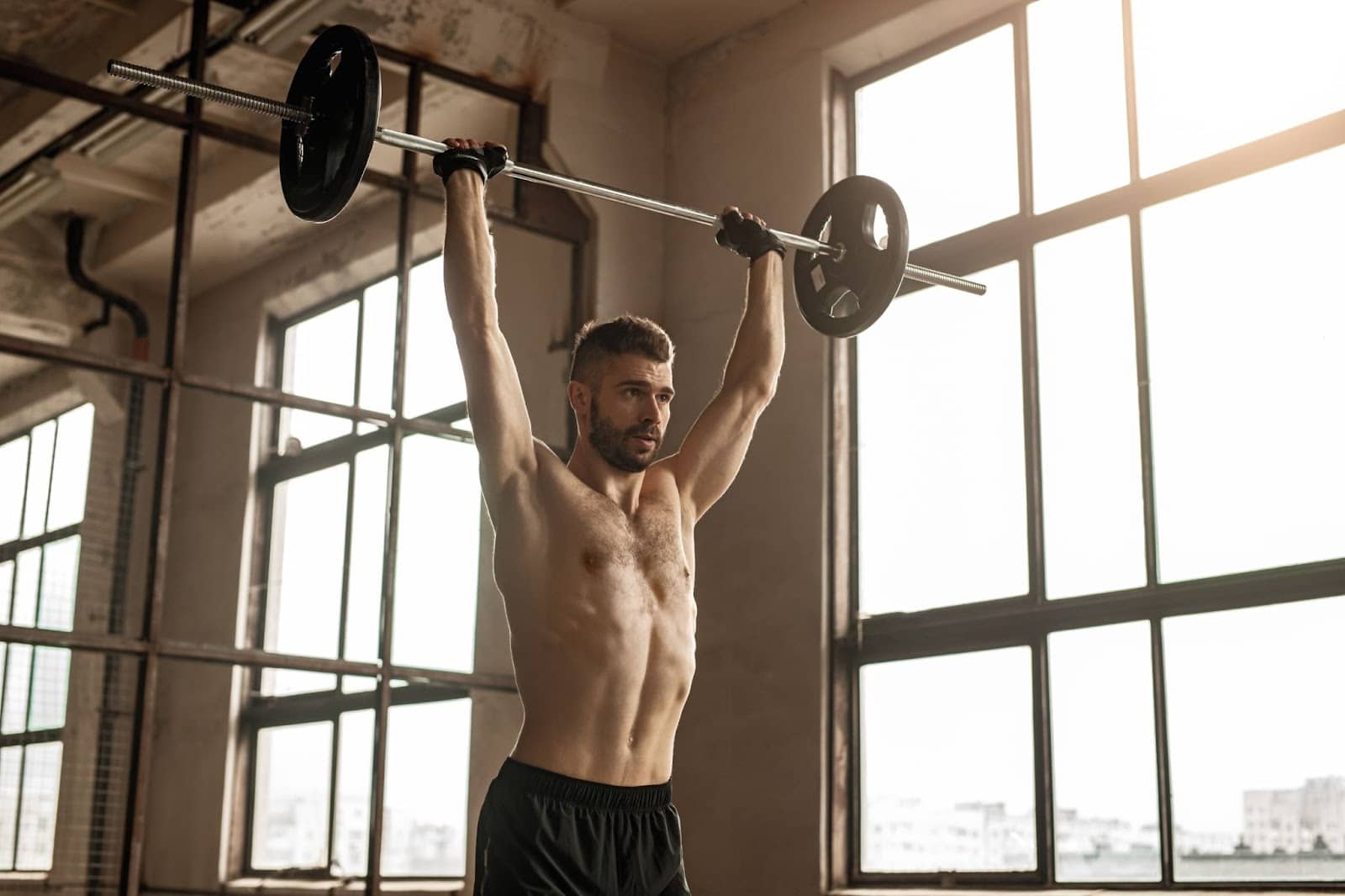 Muscular young man doing an overhead press in front of several large windows.