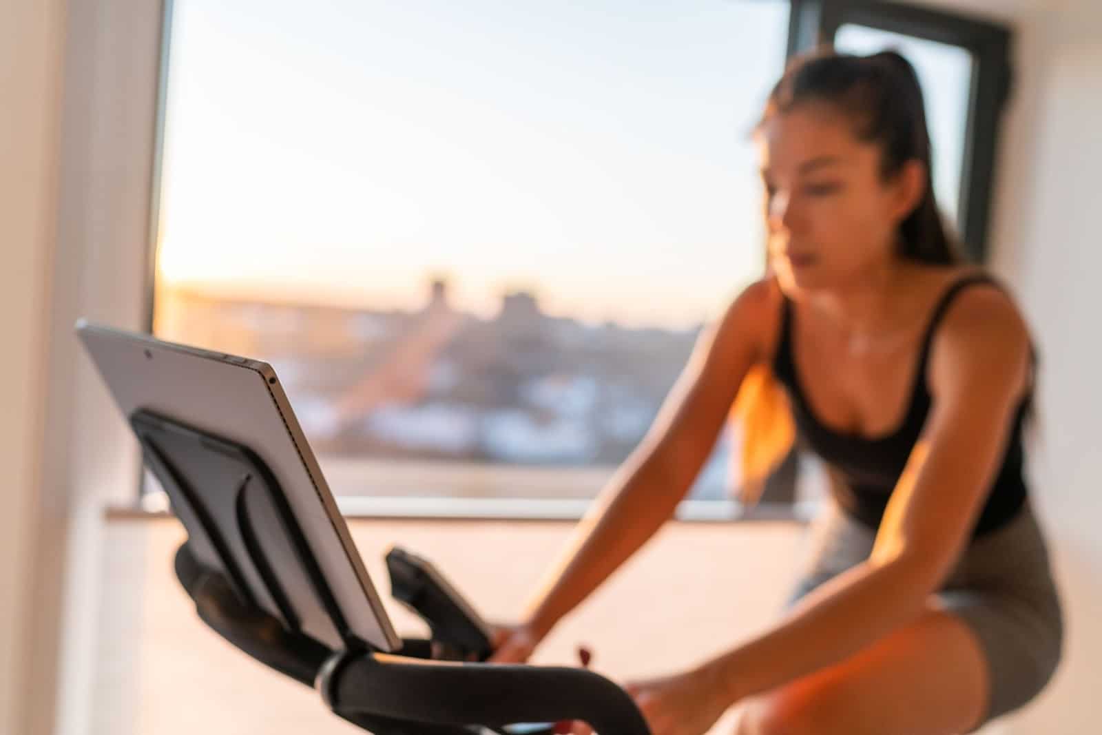 Young fit woman on home spin bike, taking a class on her laptop.