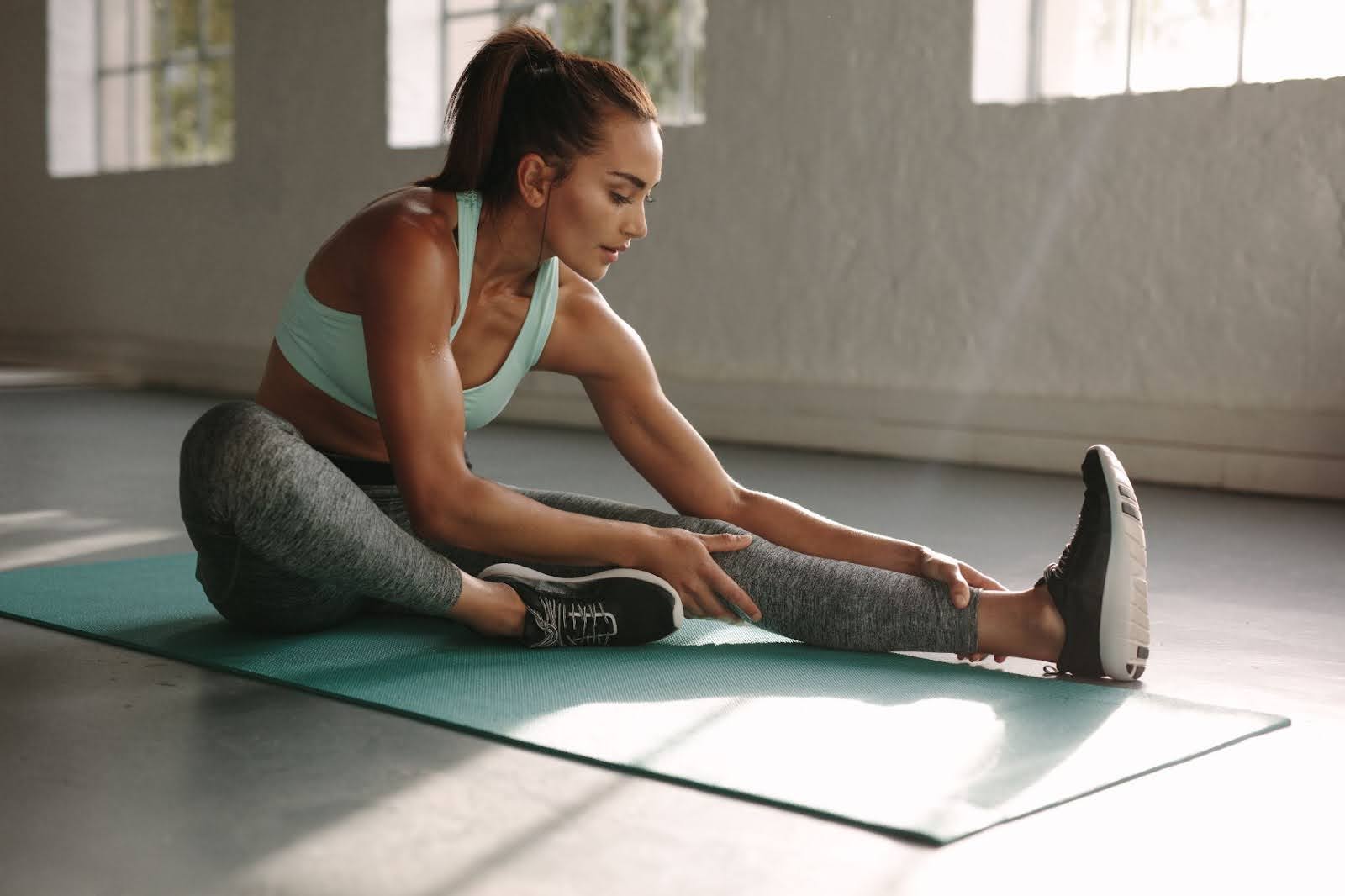 Fit woman in a green top and gray pants stretching her legs in a gym